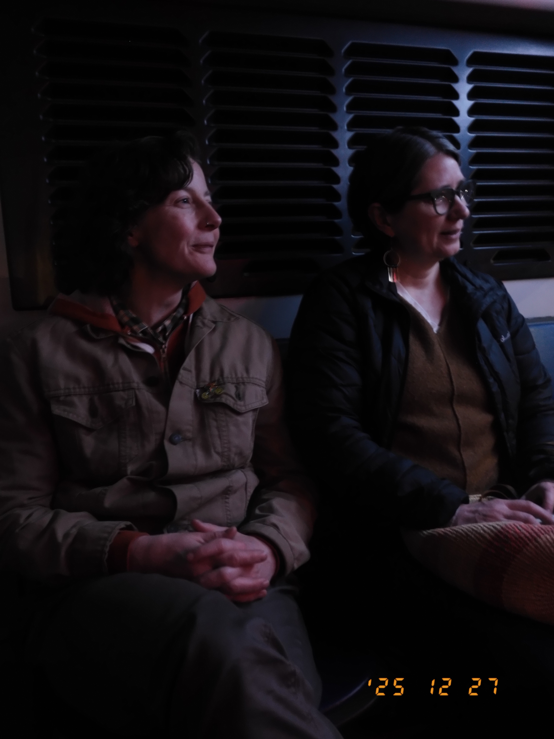 Auto-generated description: Two people are sitting closely together in a dimly lit setting with a patterned background behind them.