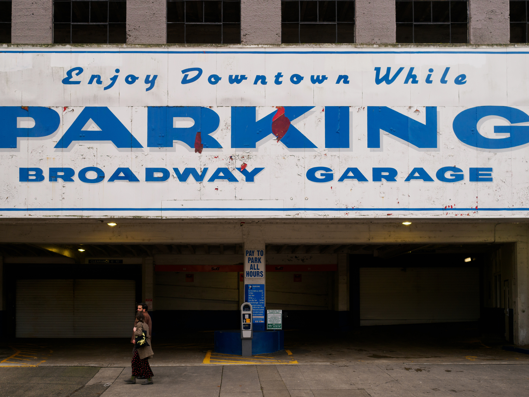 A large sign for the Broadway Garage advertising parking in downtown is displayed above a parking garage entrance, with two people walking below.
