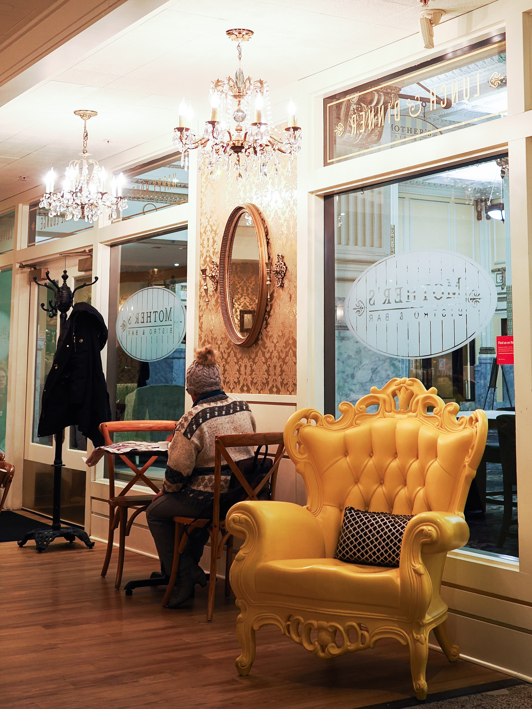 A cozy café interior features elegant chandeliers, a yellow tufted armchair, and a person sitting at a table by the window.
