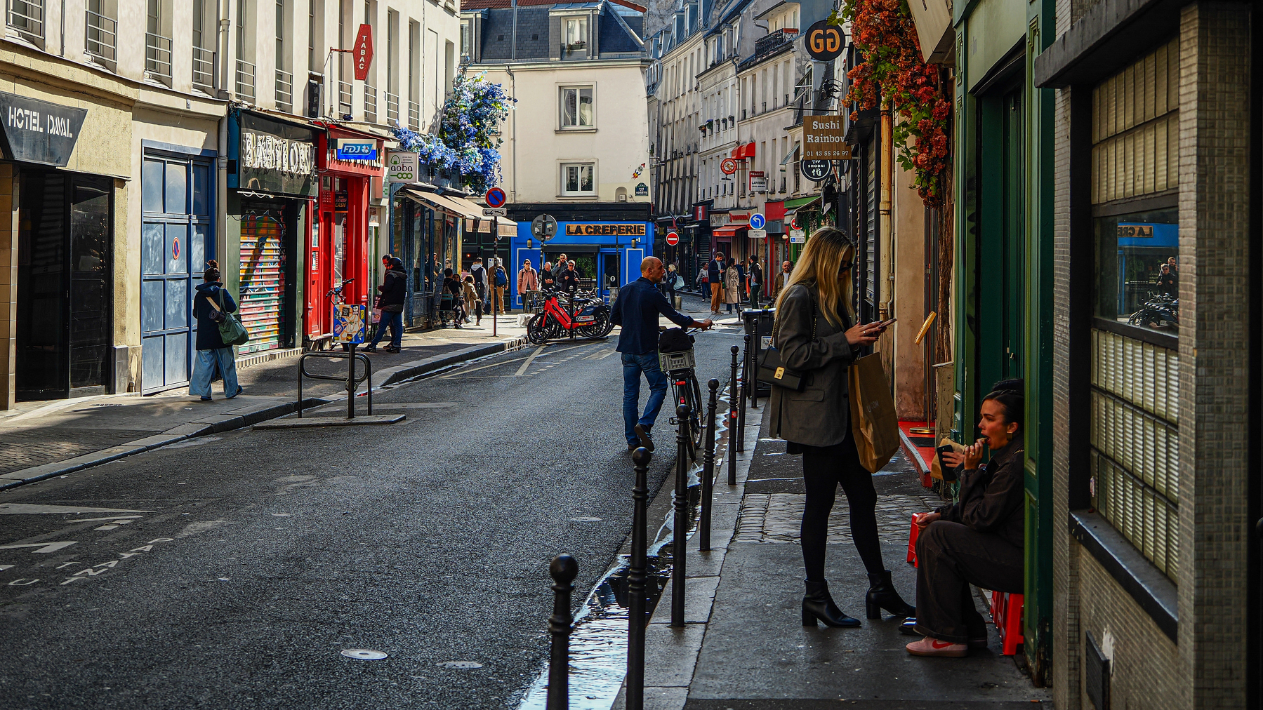 A quiet street scene in a European city features people walking, a few sitting, and colorful buildings lining the street.