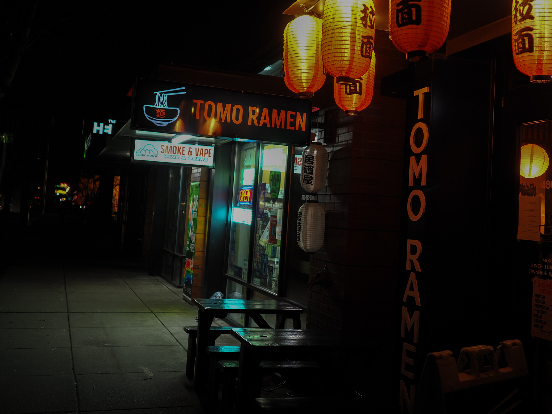 A dimly lit street features a storefront for Tomo Ramen with illuminated Japanese lanterns and adjacent vape shop signage.