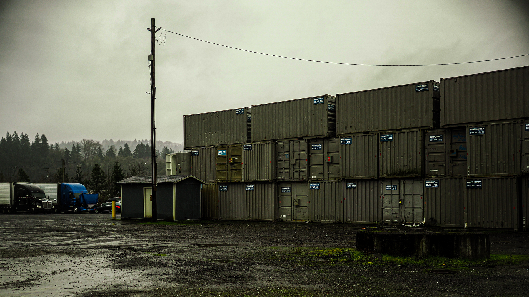 Stacked shipping containers are arranged outdoors near a small utility building on a cloudy day.
