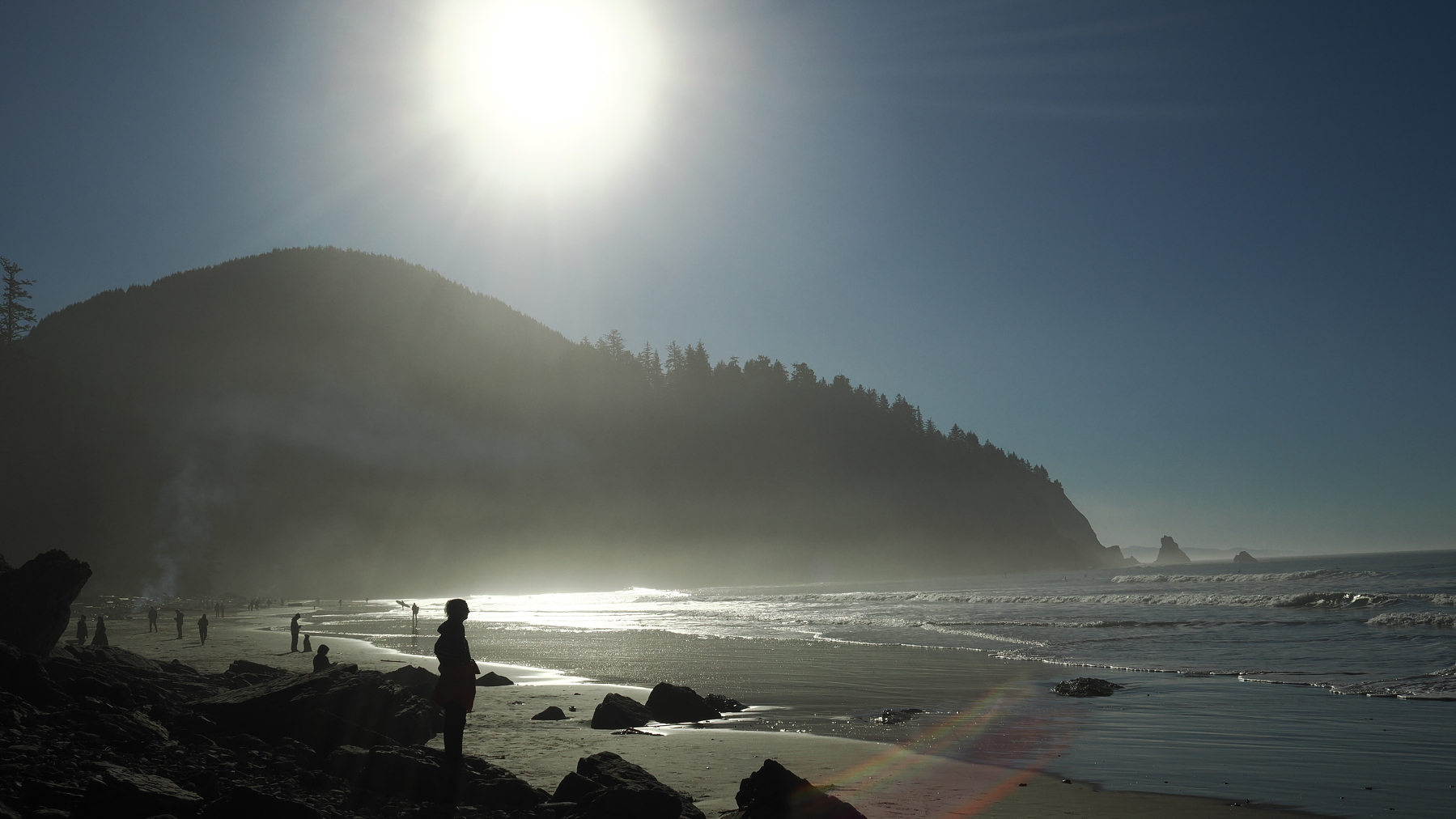 A serene beach scene features silhouettes of people along the shoreline, a bright sun in the sky, and a tree-covered hillside.