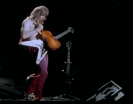Nigel Tufnel plays an electric guitar with a violin  stage next to another guitar standing on a stand.