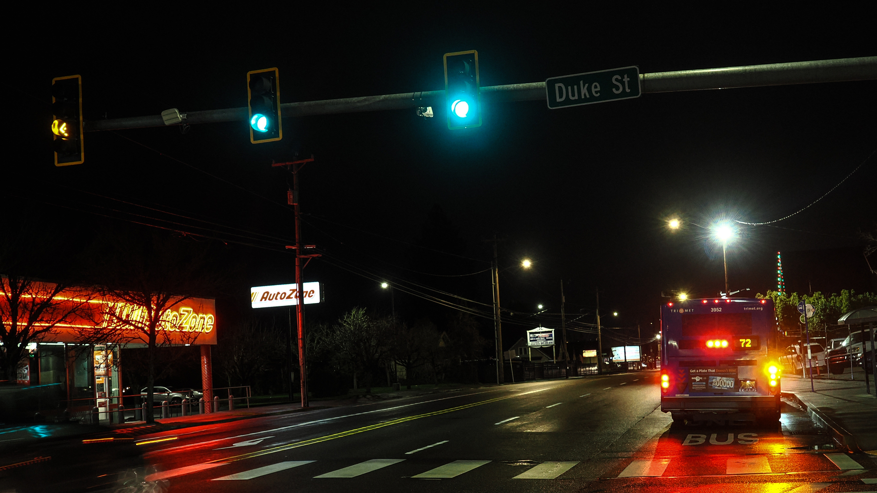 A city street at night shows a bus stopped under traffic lights near a neon-lit AutoZone.