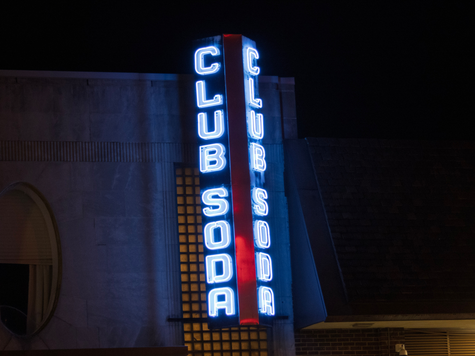 A neon sign displaying the words CLUB SODA is illuminated against a dark night backdrop.