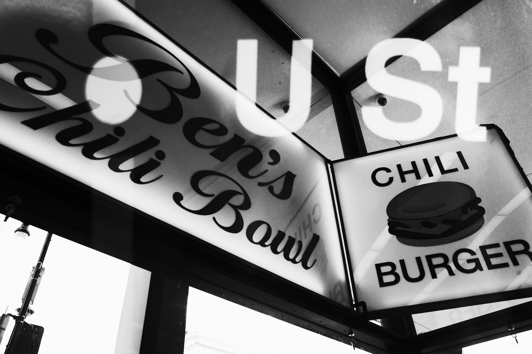 A black and white photo shows signs for Ben's Chili Bowl and Chili Burger with U St reflected above.