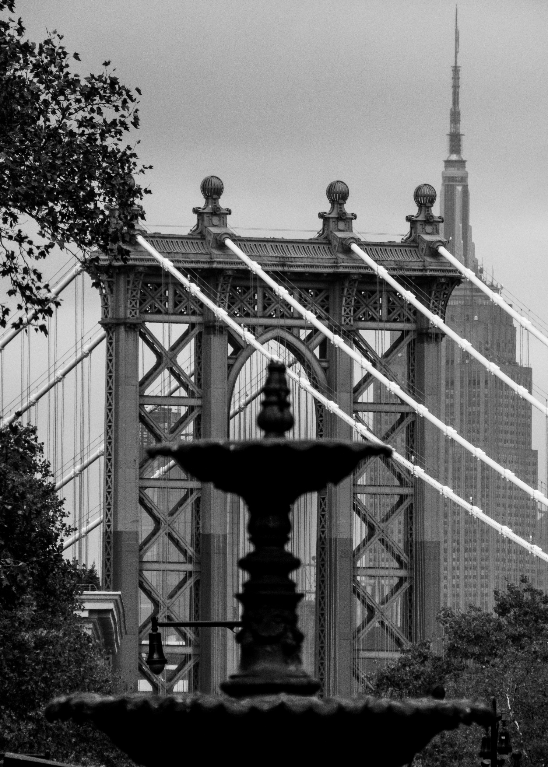 A black and white photo shows the Manhattan Bridge with the Empire State Building visible in the background and a silhouette of a fountain in the foreground.