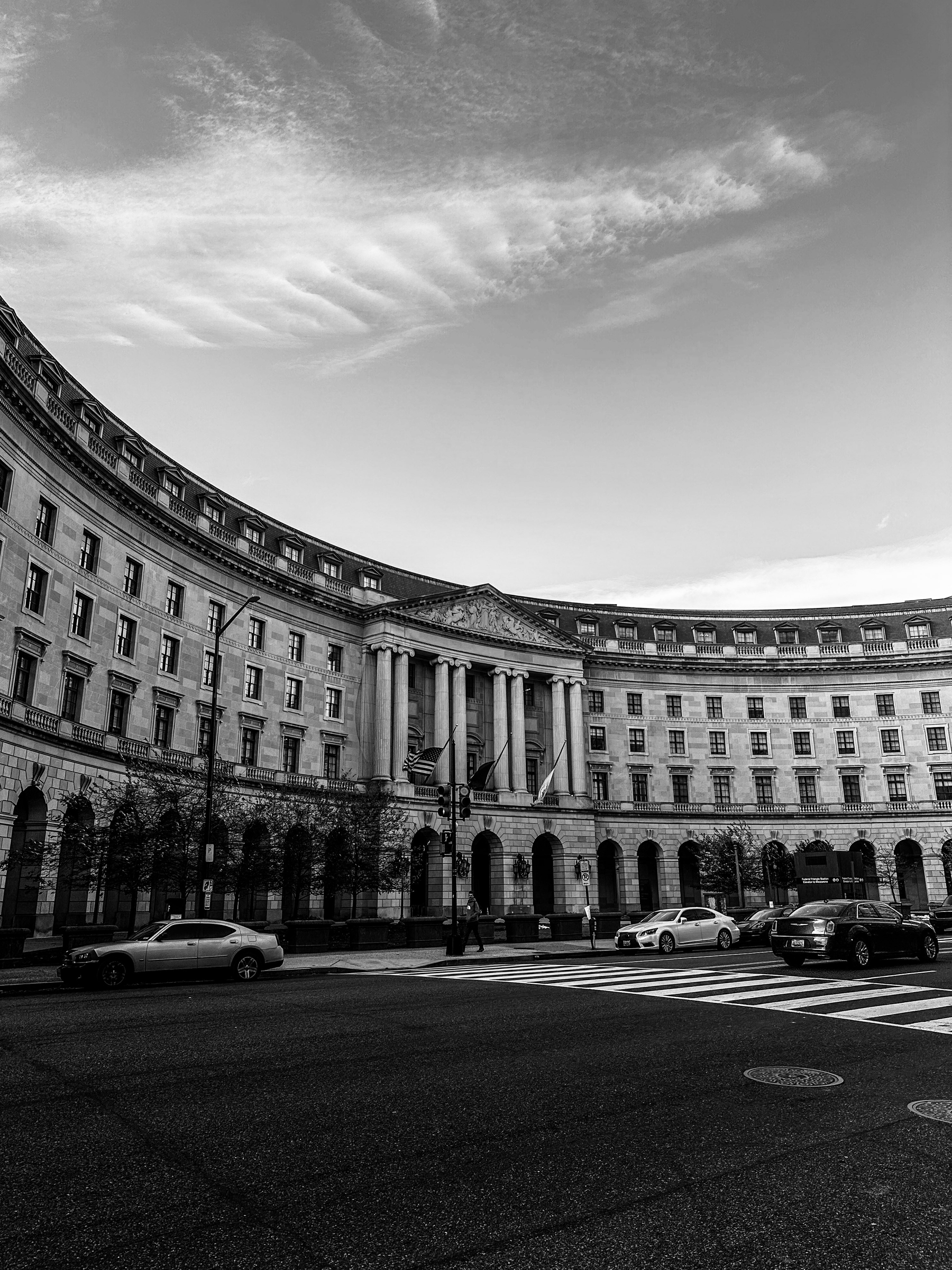 A large, curved building with classical columns and arched windows is shown in black and white with cars parked in front.