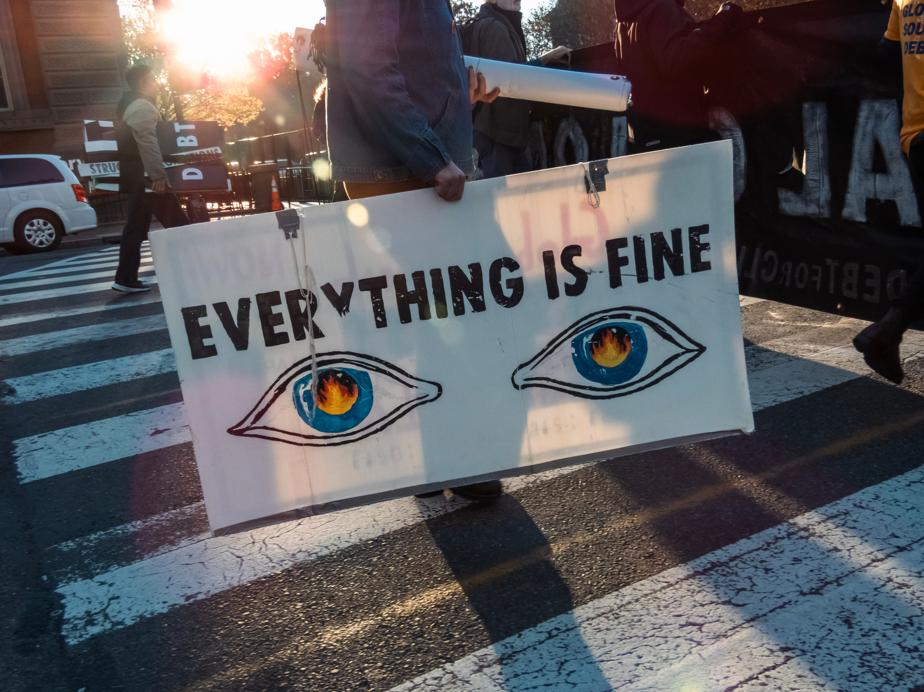 A person holds a sign with the text EVERYTHING IS FINE and two illustrated eyes, while walking on a zebra crossing during a sunlit moment.