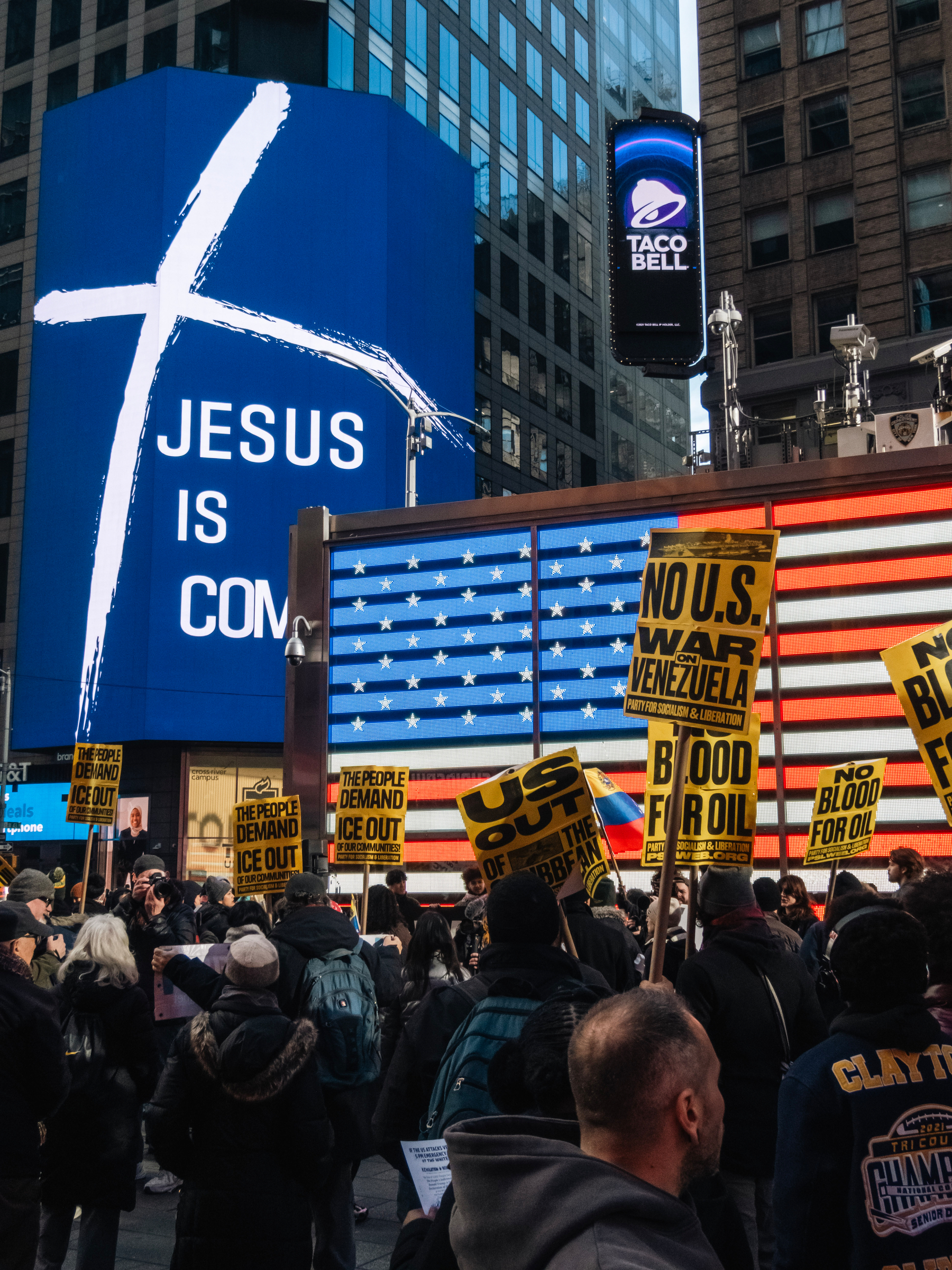 Protesters are gathered in an urban setting with anti-war signs, while large digital displays in the background show a cross with the text JESUS IS COMING and an American flag.