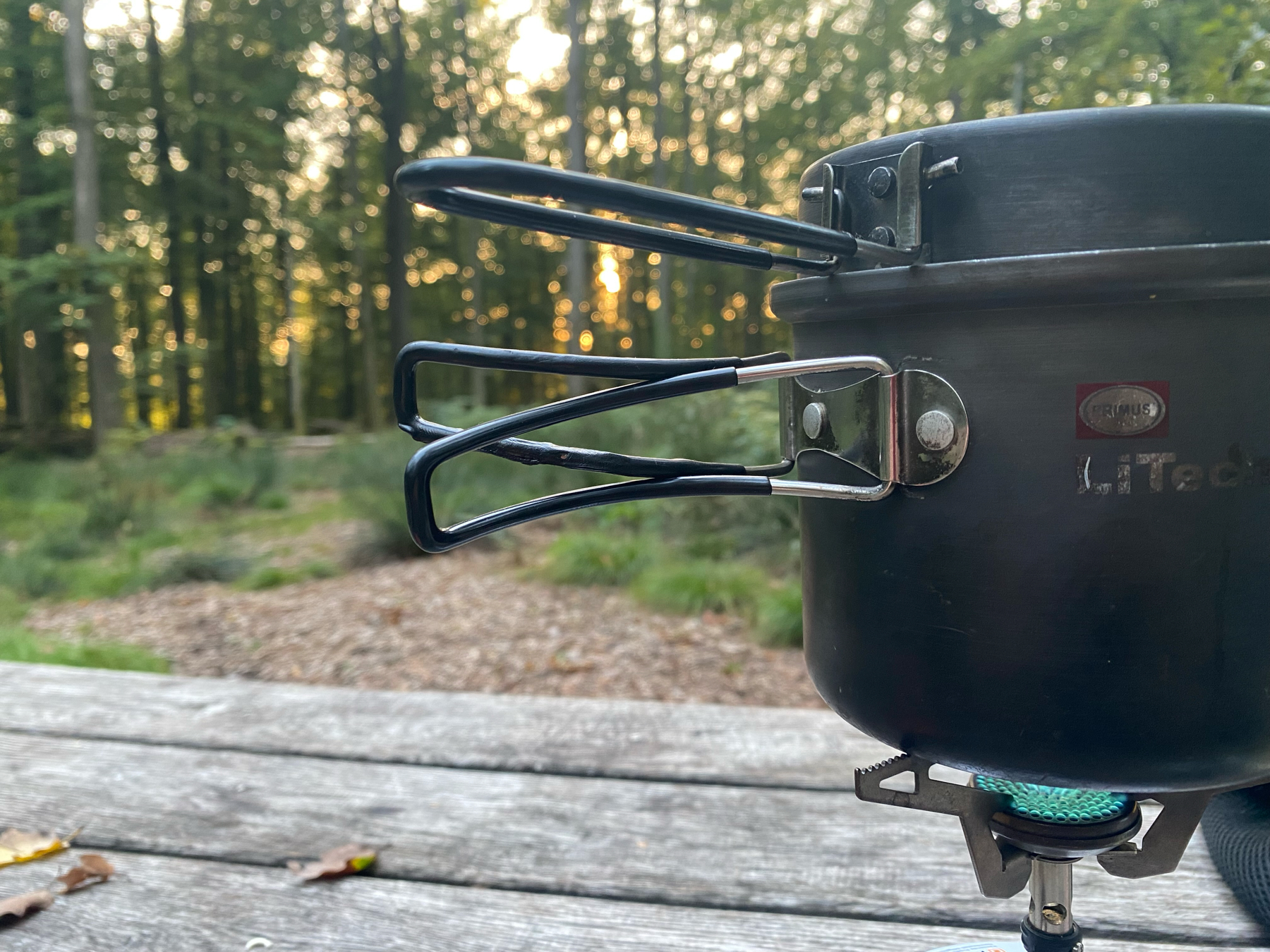 camping pot on camping stove with a blurry Forrest in the back, the low sun peeking through the trees