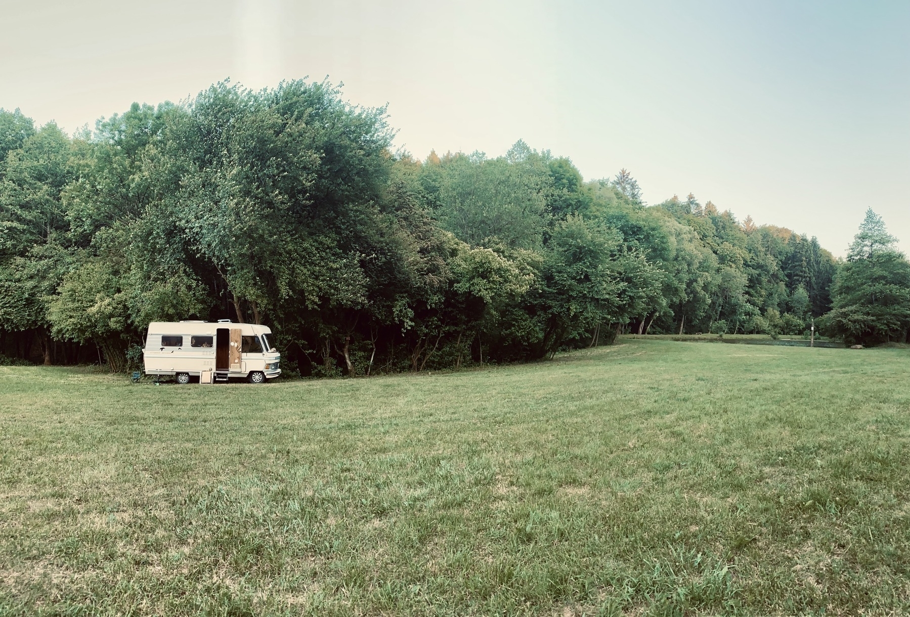 An old mobile home is parked in a meadow in front of a forest.&10;
