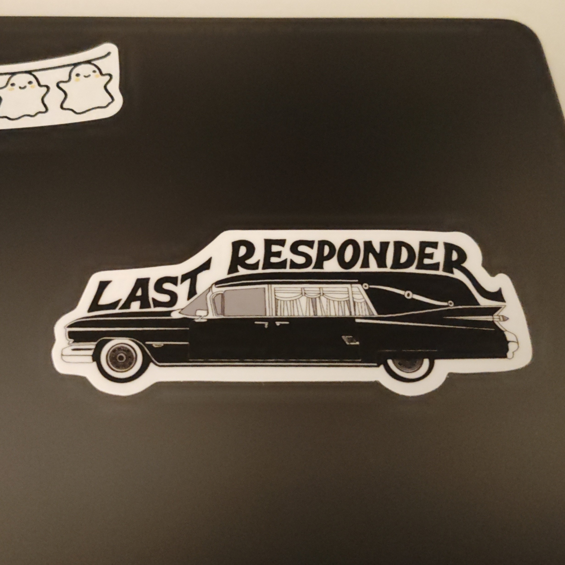 A sticker on a black laptop. The sticker has the words last responder above a hearse.