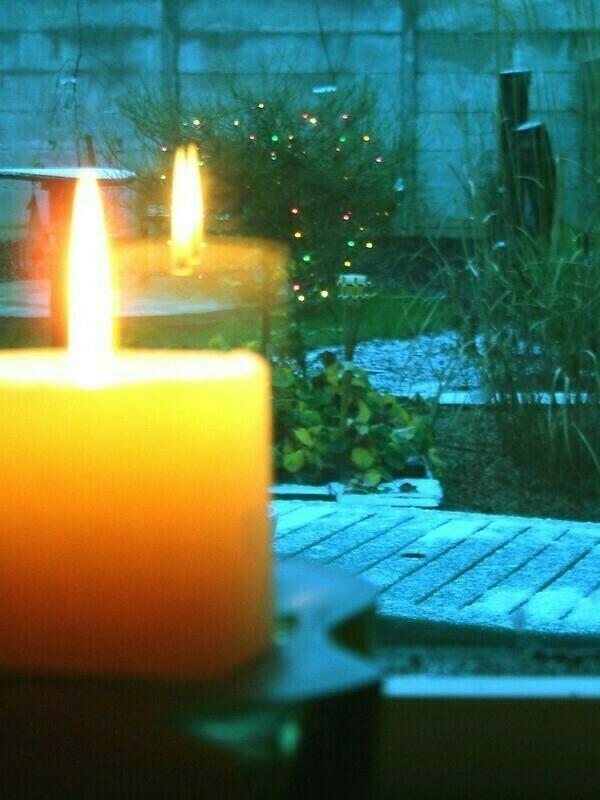 A candle is in the foreground with its flame reflected in our Garringreen window, while a garden and a bush with colored lights are visible outside.