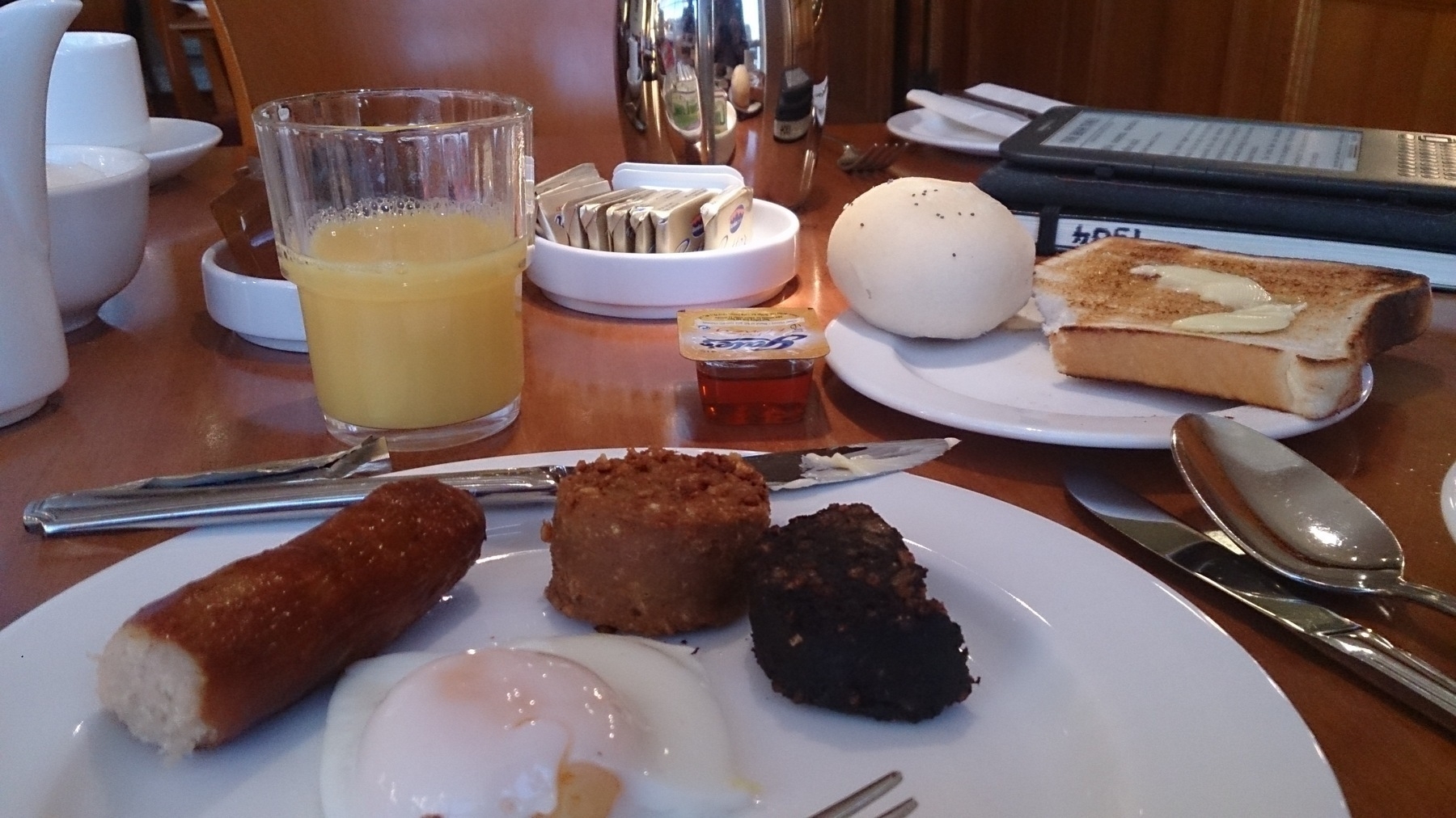 Auto-generated description: A breakfast spread featuring a plate of sausages, a poached egg, black pudding, toast with butter, an orange juice, and a bread roll on a wooden table.