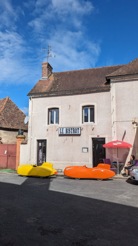 A quaint café named Le Bistrot features a cozy outdoor seating area with colorful paddle boats as decorations under a bright blue sky.