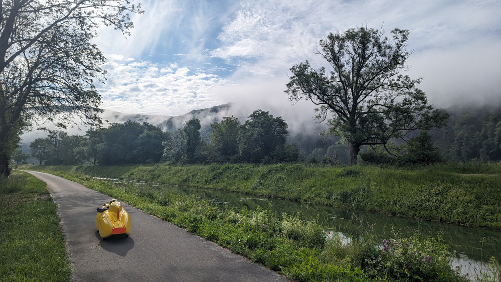 A yellow velomobile travels along a paved path beside a tranquil river, with misty hills and trees in the background.