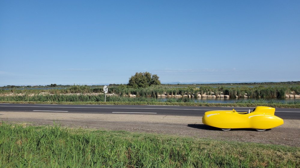 A bright yellow velomobile travels along a road bordered by grass and a scenic, expansive landscape.