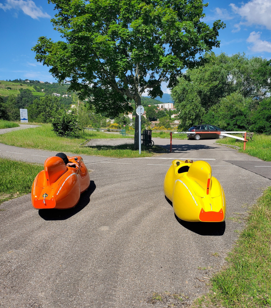 Two velomobiles, one orange and one yellow, are on a paved path surrounded by greenery with a tree and car in the background.
