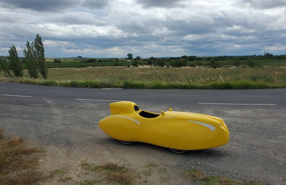 A yellow, aerodynamic velomobile is parked on the side of a rural road with a cloudy sky overhead.