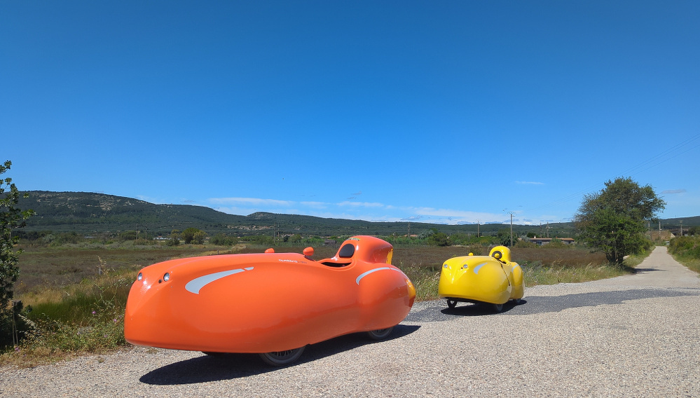 Two colorful velomobiles, one orange and one yellow, are parked on a rural road against a backdrop of blue sky and rolling hills.