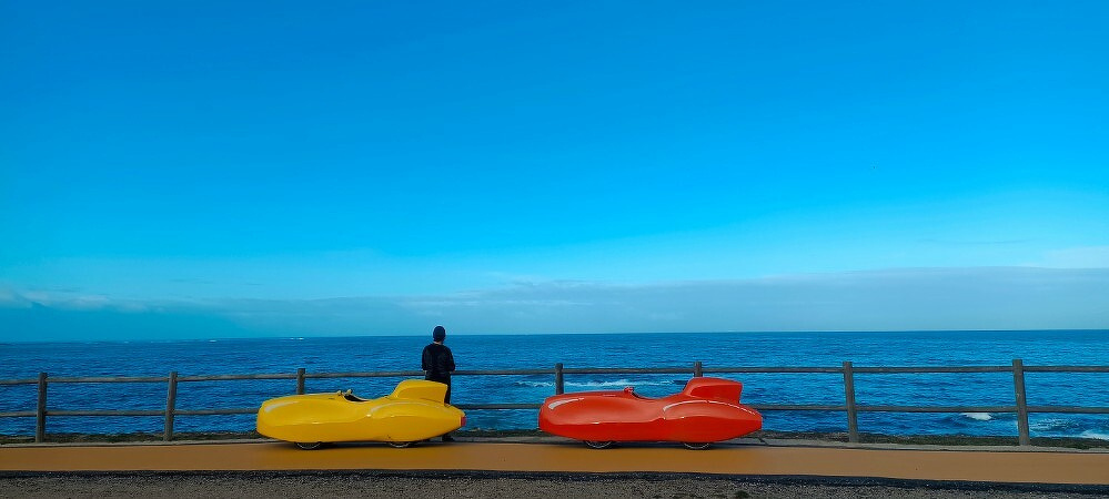 A person stands on a boardwalk overlooking the ocean with colorful, abstract benches in the foreground.