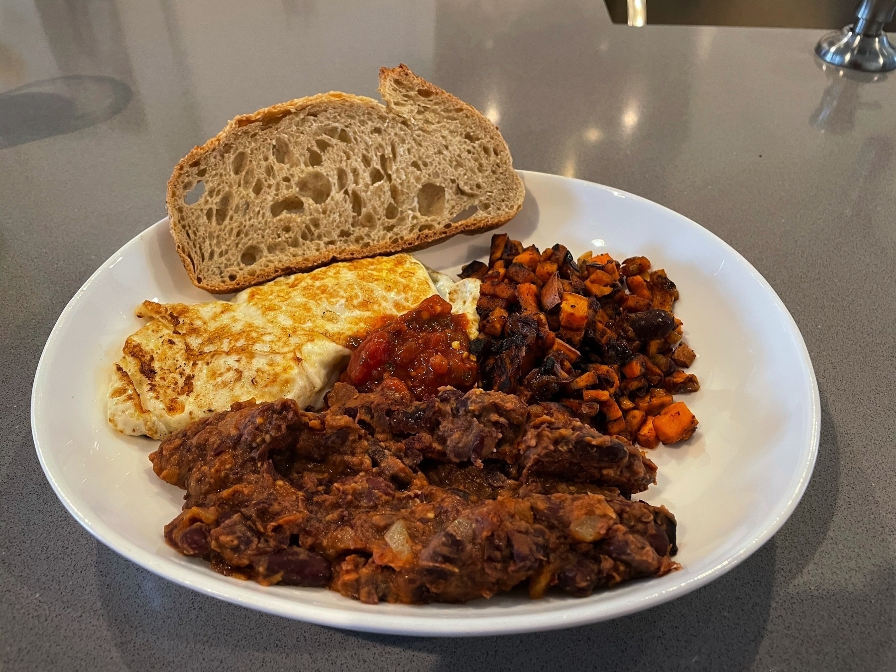 Breakfast plate with two eggs, sweet potato hash, beans, and a slice of sourdough.