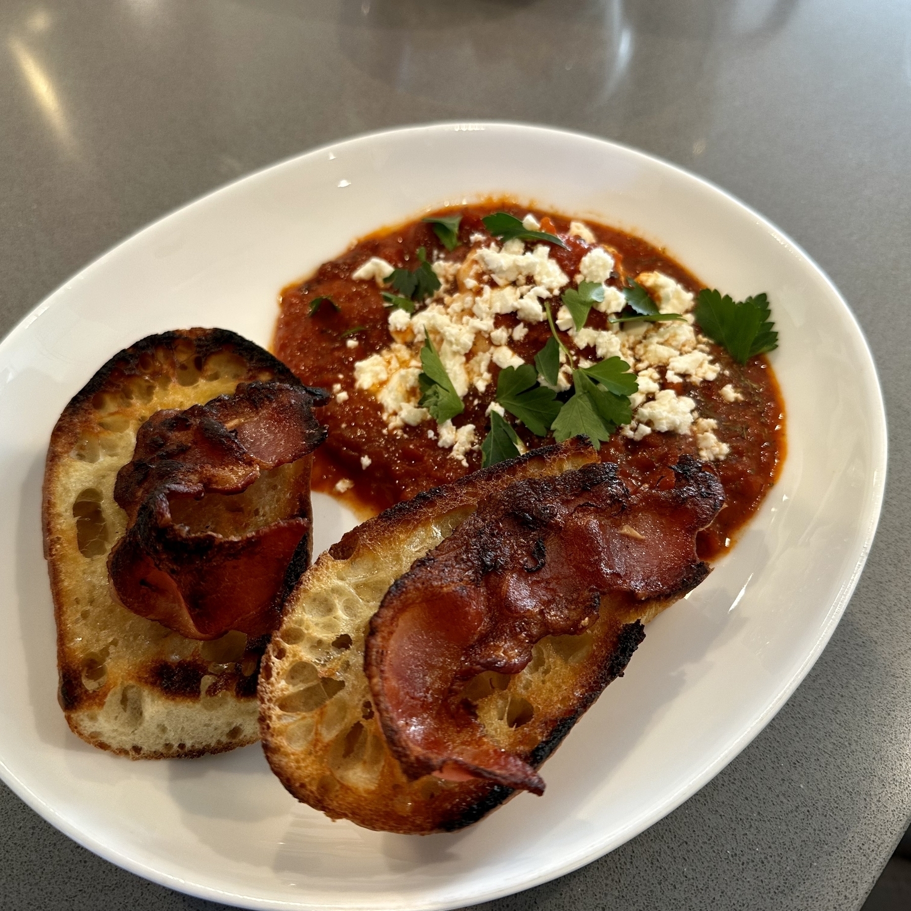 A toasted baguette with two strips of bacon adjacent to a pool of crushed, cooked tomatoes with a poached egg, feta, and fresh parsley.