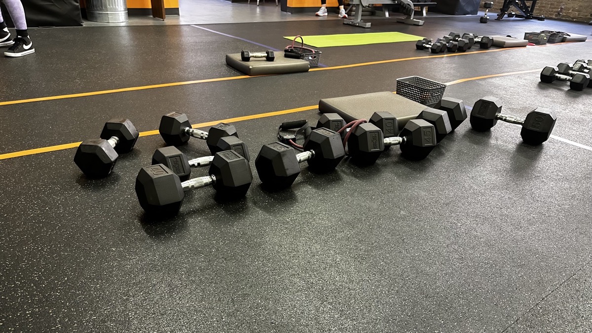 A pile of dumbbells on a rubber floor that has boxes taped out at a gym.