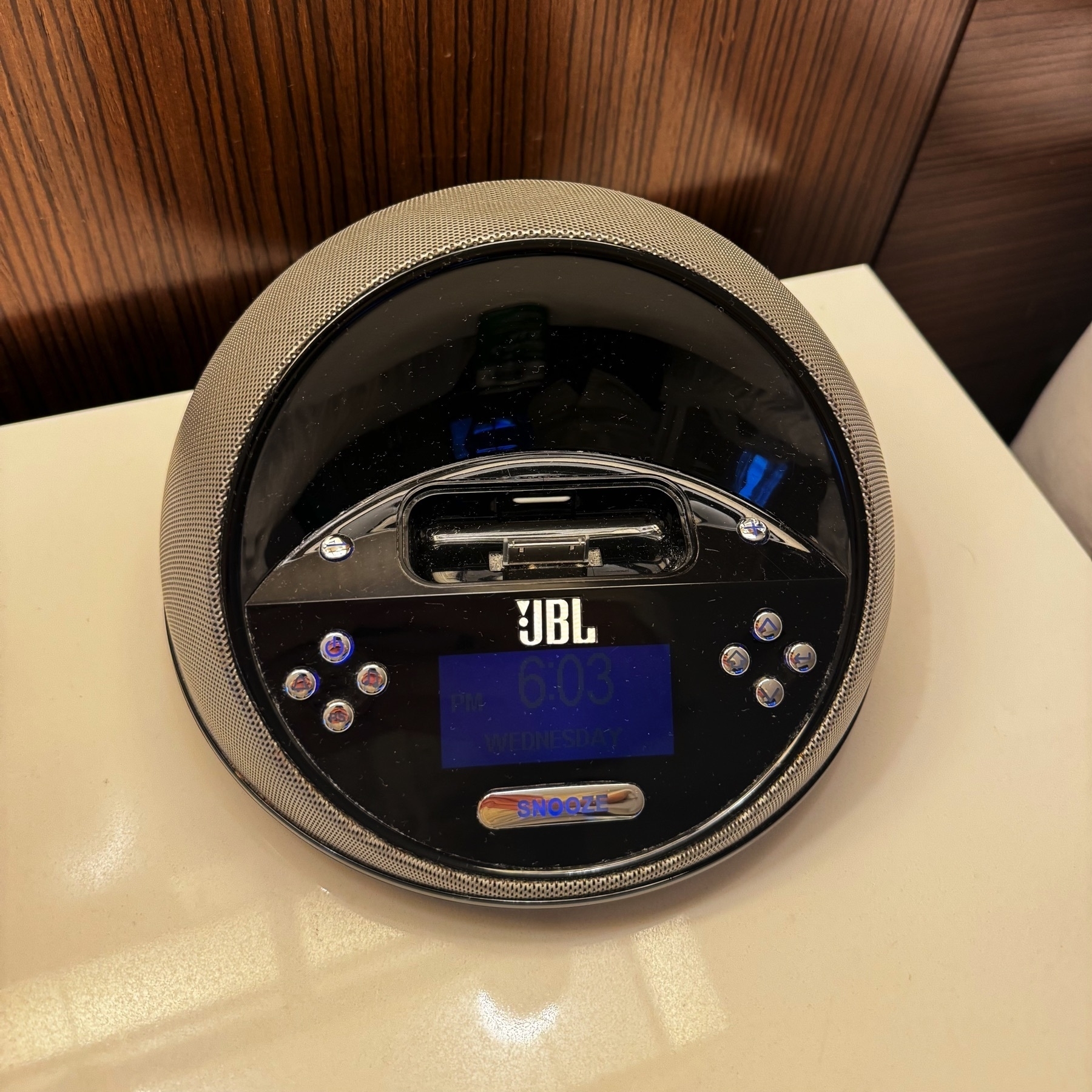 A JBL alarm clock and speaker with an iPod 30-pin connector. It’s circular on a white table with a glossy wood panel in the background.