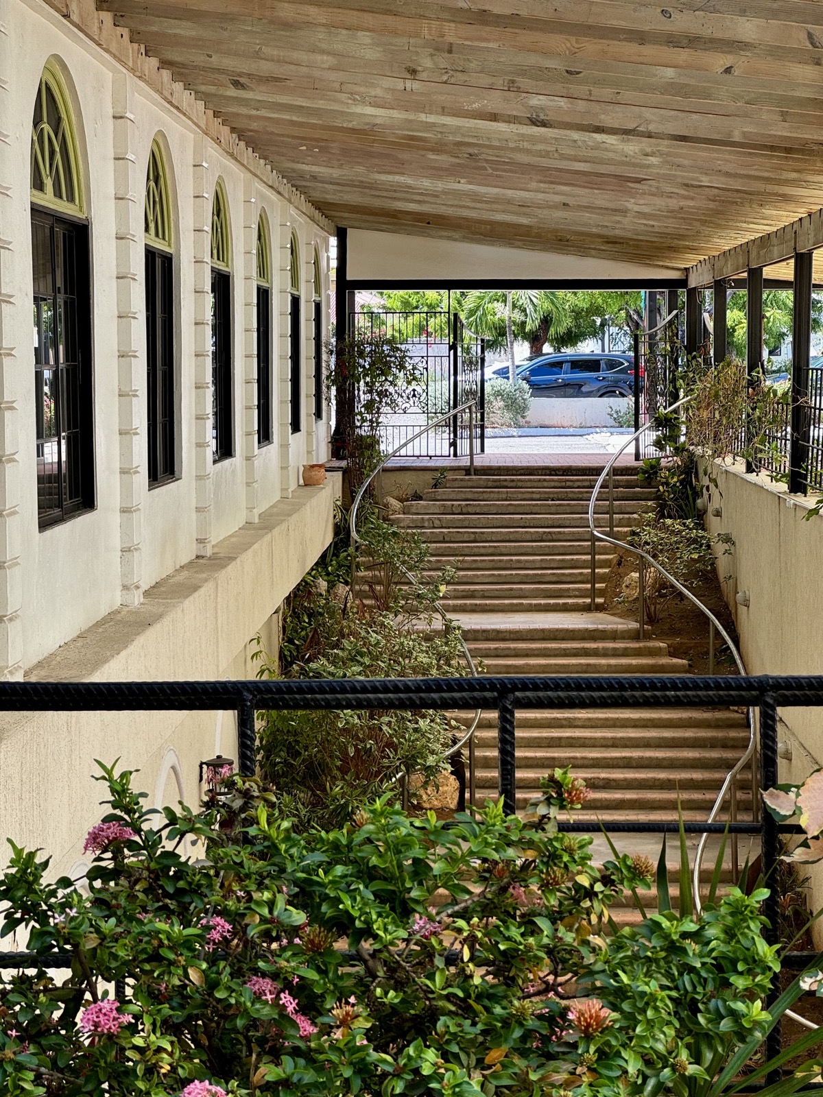 Green flowering bushes in the foreground in front a of a staircase with winding metal rails and windows with arches in a beige, stone building to the left.
