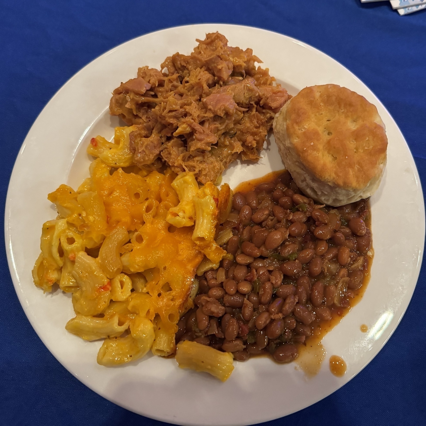 A plate of macaroni and cheese, pulled pork, baked beans, and a biscuit on a blue table cloth. The joke is that all the food is yellow, white, or brown.