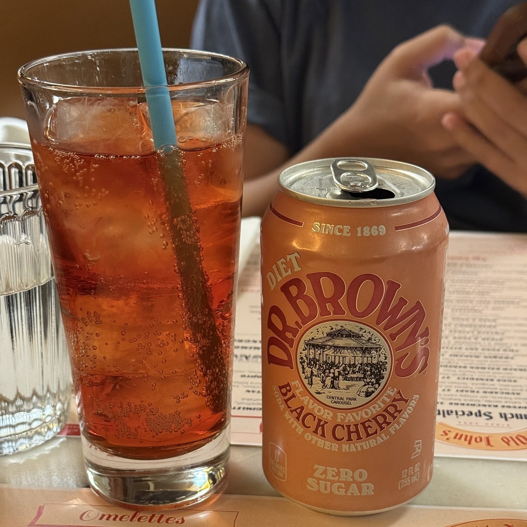 A can of Diet Dr. Brown's Black Cherry soda is placed on a table next to a glass with a straw, with a person using a phone in the background.