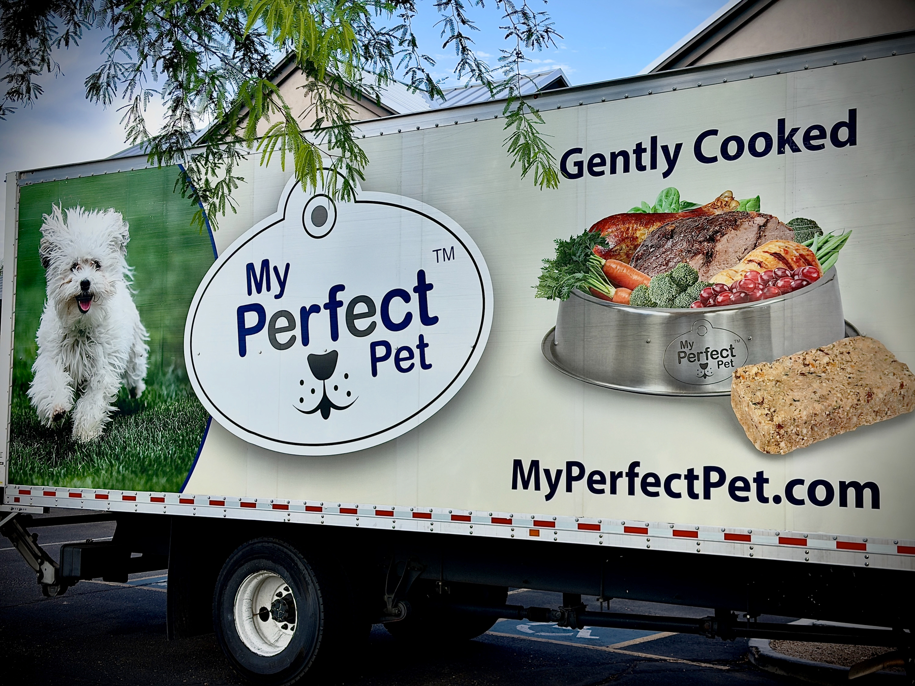 Auto-generated description: A truck features an advertisement for My Perfect Pet, showcasing a happy white dog, a pet food bowl filled with cooked ingredients, and the slogan Gently Cooked.