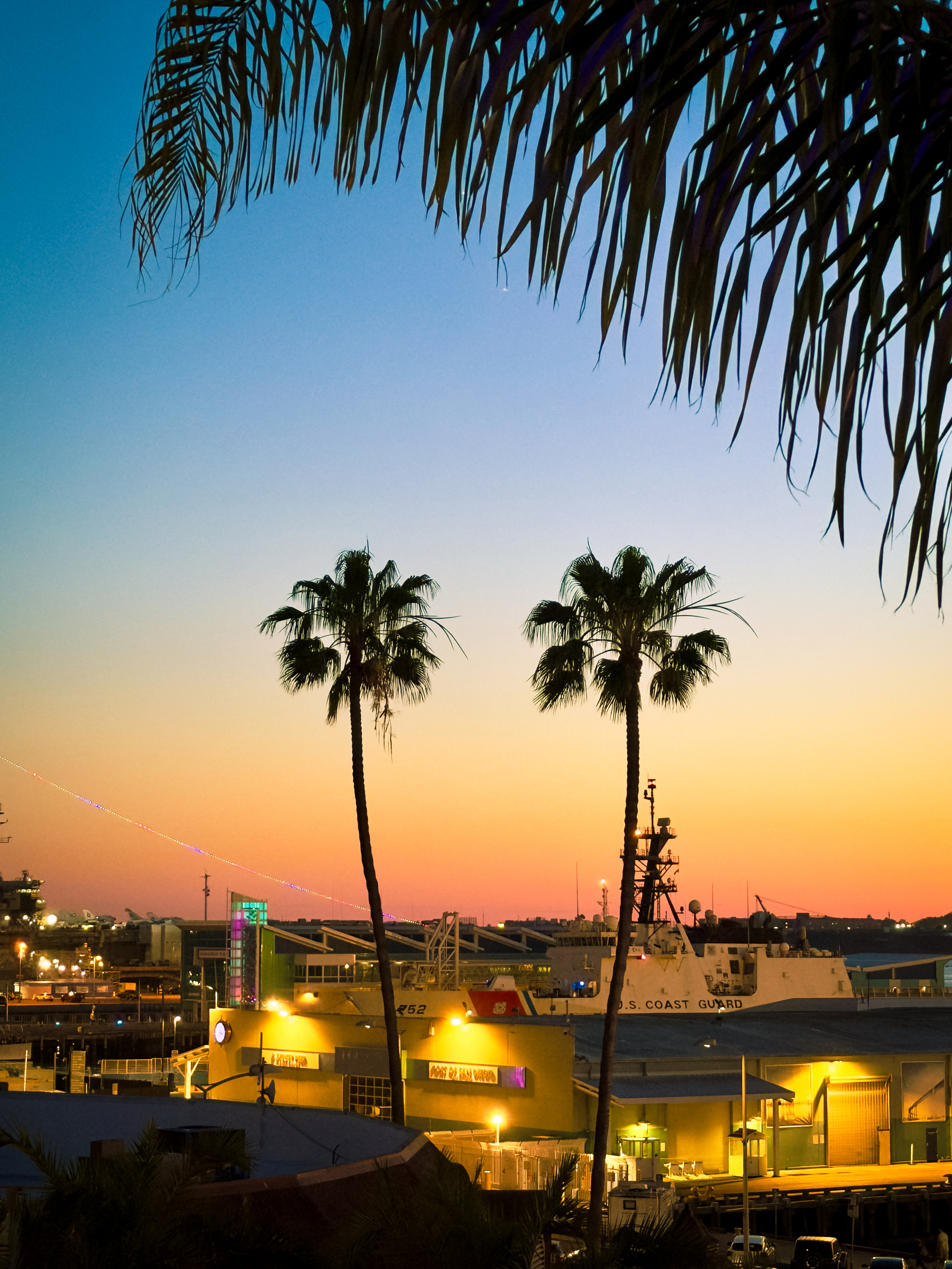 Auto-generated description: Palm trees are silhouetted against a colorful sunset sky, overlooking a coastal area with buildings and a Coast Guard ship.