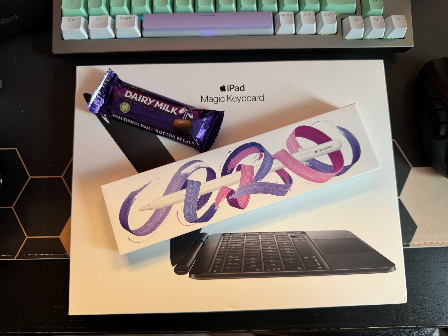 A new Apple Pencil box on a new Magic Keyboard box, with a dairy milk bar for reference. The Apple Pencil box has pink and purple doodle brush strokes on it.