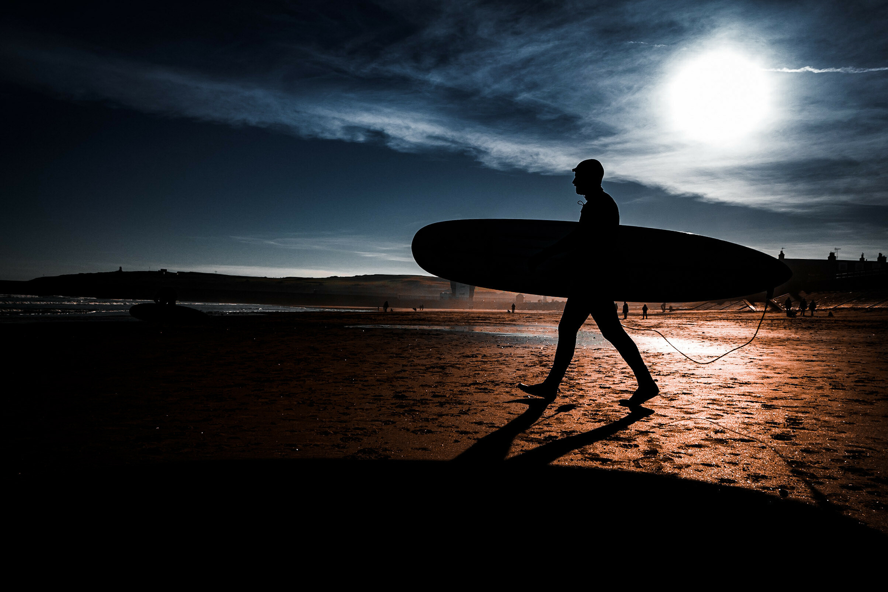 Silhouette of person carrying surfboard walking across wet beach at sunset with dramatic cloudy sky and bright sun breaking through.​​​​​​​​​​​​​​​​