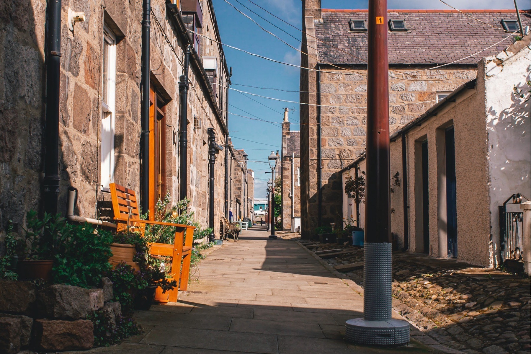 Narrow lane in Fittie, Aberdeen, between stone cottages with orange bench and planters, overhead wires crossing blue sky, leading towards water on a summer day.​​​​​​​​​​​​​​​​