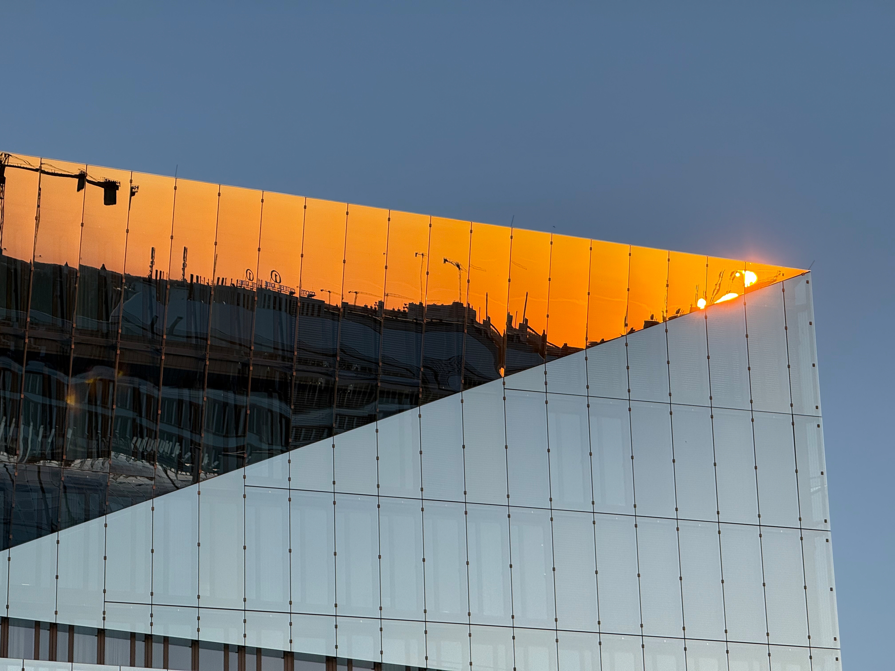 A modern glass cube shaped building reflects the sunset and nearby structures, creating an angular contrast between warm and cool tones.
