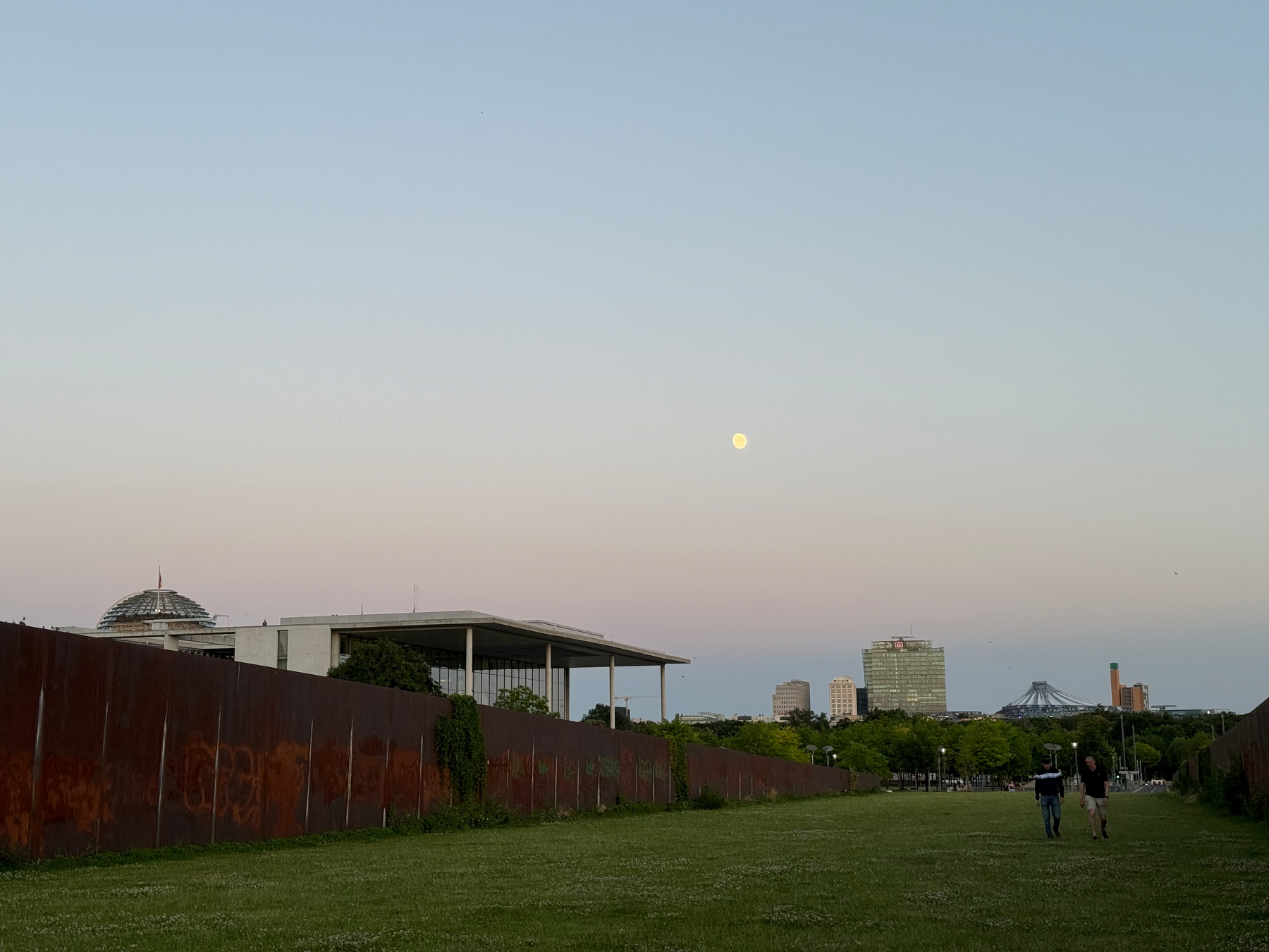 A wide grassy pathway flanked by rusty metal walls leads toward the German capitol government district under a clear sky with a visible moon, where two people are walking on the right side.
