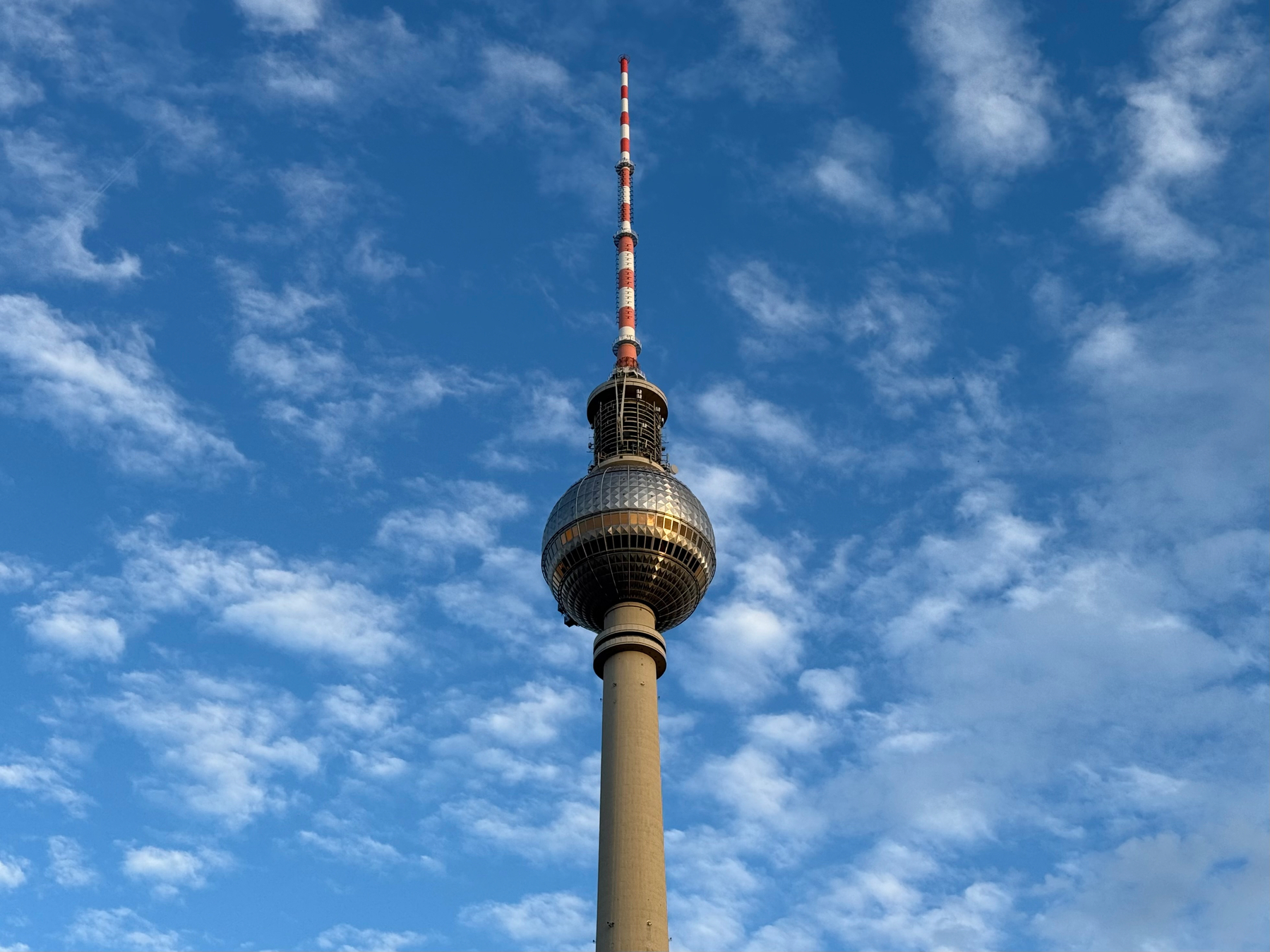 A tall tv tower with a spherical observation deck and an antenna stands against a partly cloudy blue sky.