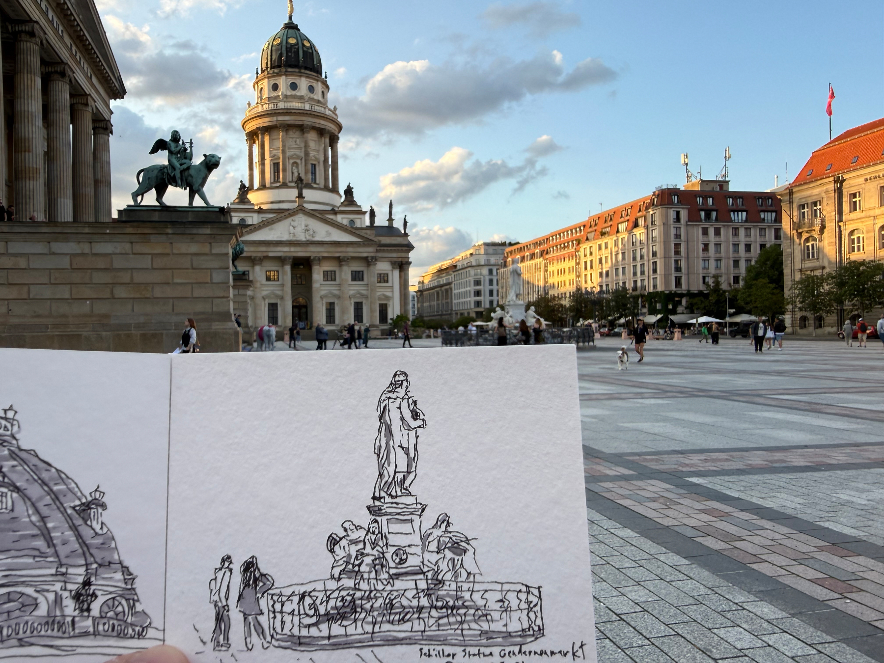 A hand-drawn sketch of a statue is held up in front of a large square with historic buildings including the steps of the Concert House and the domed French church in the background.