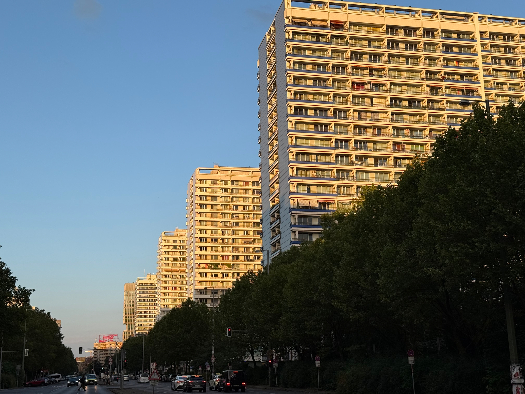 Tall wide apartment buildings line a tree-lined street under a clear blue sky during golden hour.