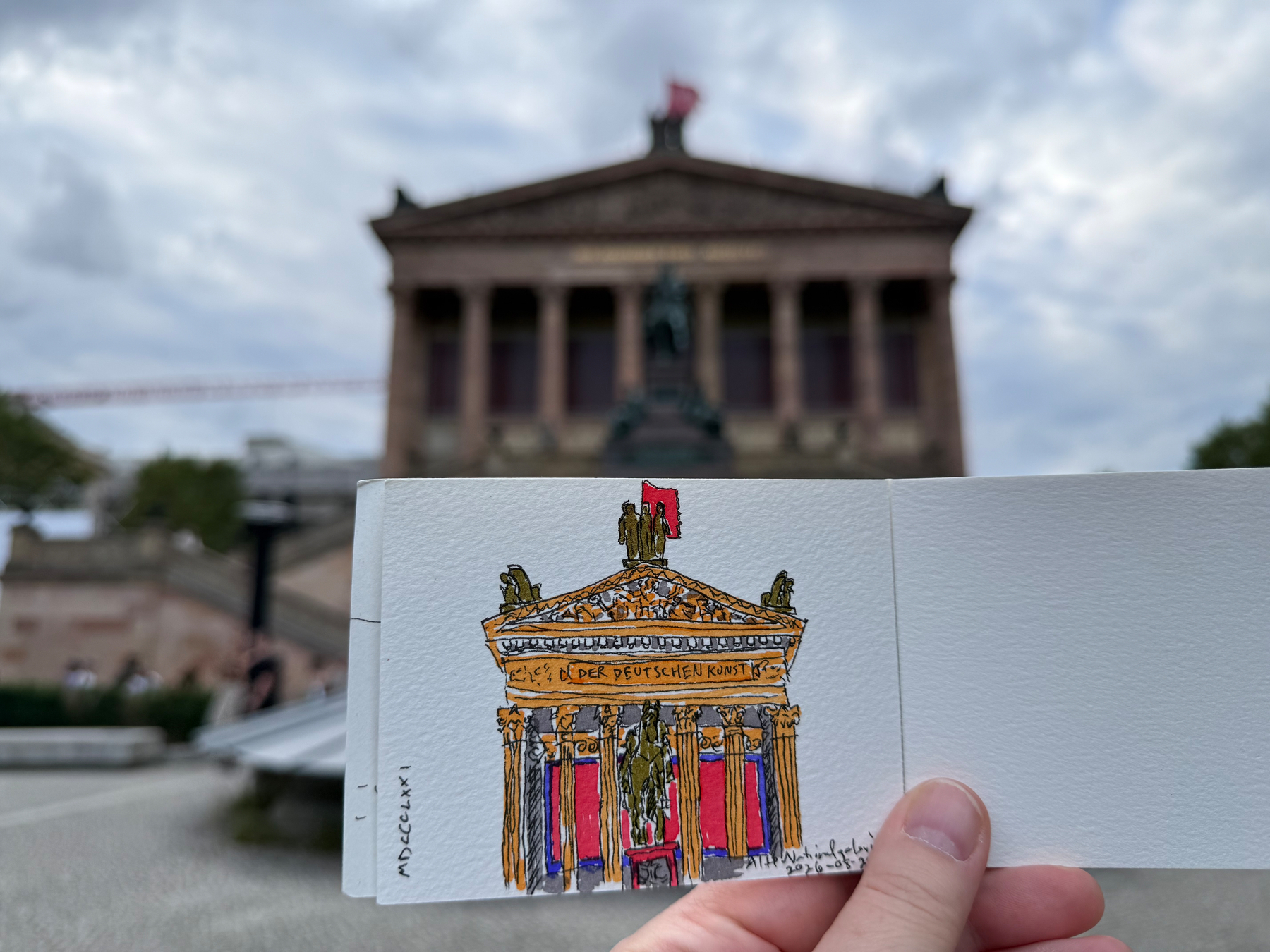 A hand holds a sketchbook with a colorful drawing of a classical museum building, matching the blurred architectural structure in the background.
