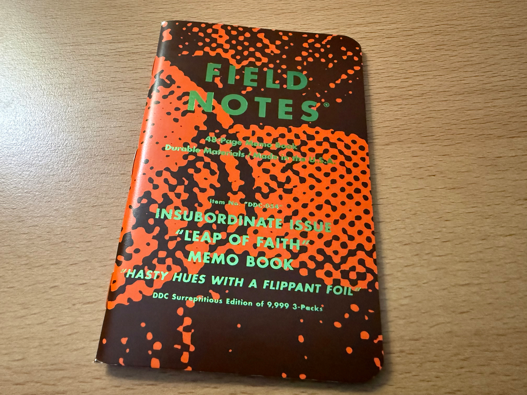 A Field Notes memo book with a brown and orange cover featuring green foil text and a bold spotted pattern sits on a desktop.
