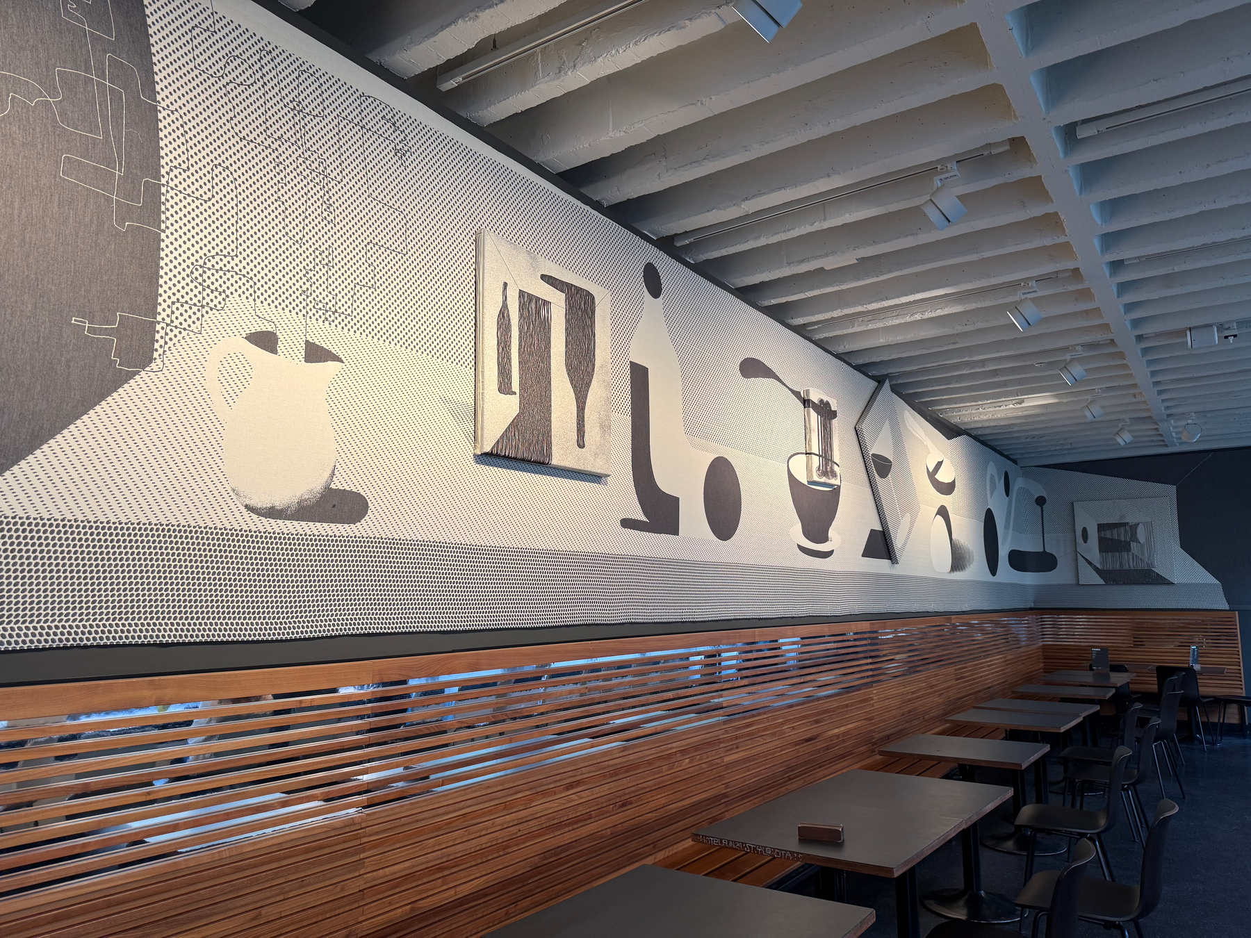 A modern, minimalist café interior features wooden seating and tables beneath a long, monochromatic woven wall mural of abstract still life of a lunch.