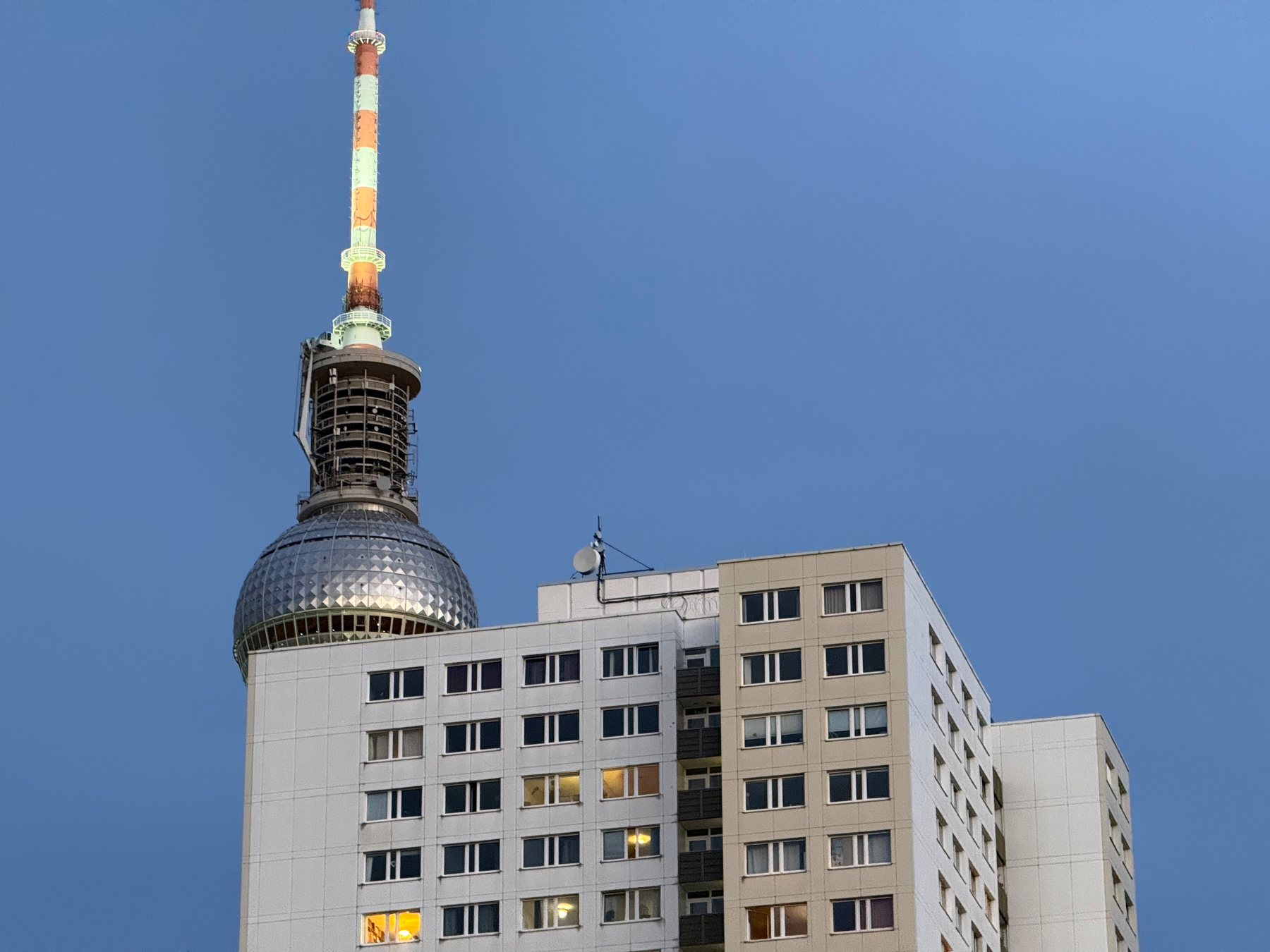 A tall spherical tv tower with an orange and white striped antenna stands behind a white residential building under a clear dusk blue sky.