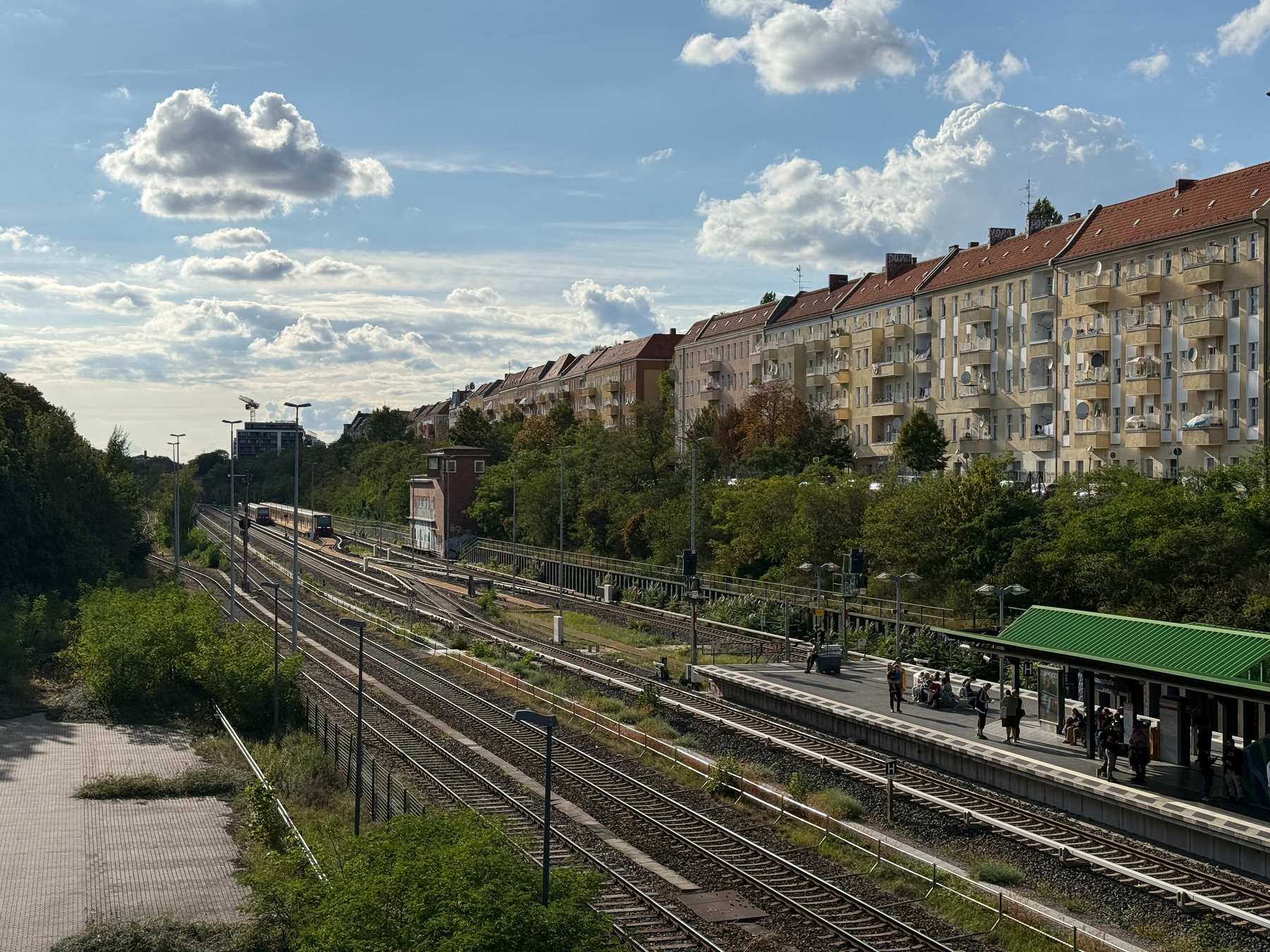 An S-Bahn station with multiple tracks and a platform where people are waiting, surrounded by trees and residential buildings under a partly cloudy sky.