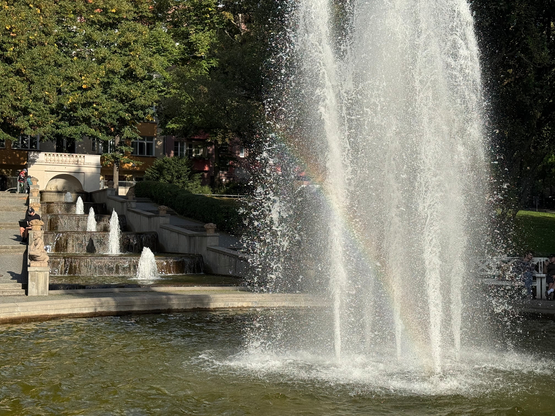 A large water fountain sprays high into the air with a visible rainbow, surrounded by greenery and stone steps with smaller cascade fountains.